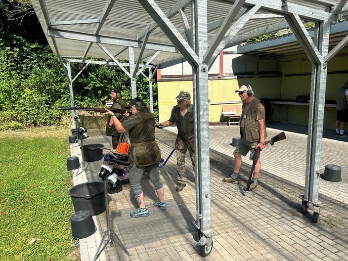 Drei Personen schießen mit Schrotflinten auf einem Schießstand im Freien. Eine Person zielt, während andere Hand lüftet. Im Hintergrund sind Bäume und ein überdachter Bereich zu sehen.