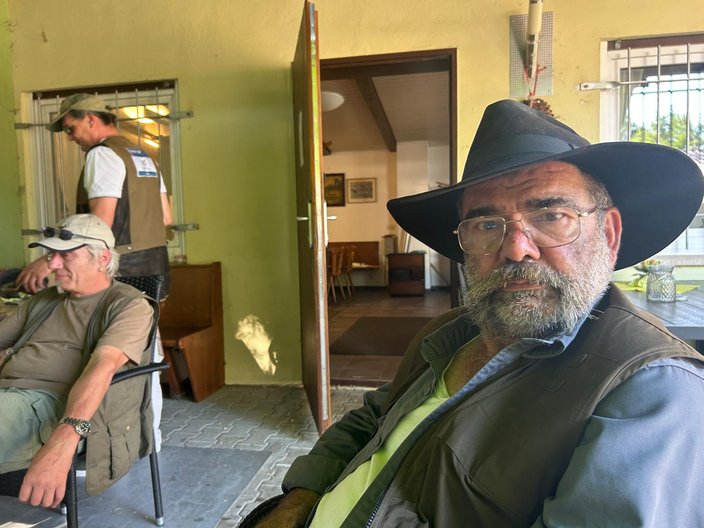 Ein Mann mit Hut sitzt und schaut in die Kamera, während ein anderer Mann vorbeigeht. Der Raum hat eine geöffnete Tür, durch die ein möblierter Innenbereich sichtbar ist.