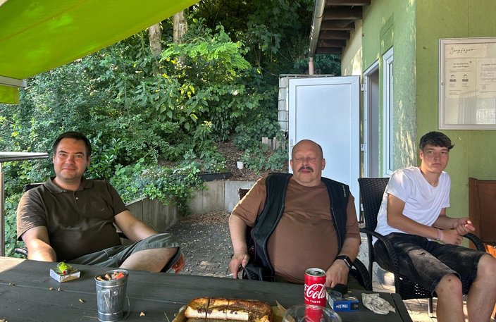 Drei Männer sitzen auf einer Terrasse. Einer trinkt Cola und ein anderer hat die Augen geschlossen. Im Hintergrund sind Grünpflanzen und eine offene Tür sichtbar.