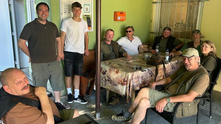 Eine Gruppe von neun Personen sitzt und steht entspannt um einen Tisch mit Tischdecke. Sie befinden sich in einem überdachten Außenbereich, erkennbar an Wänden und Tür. Im Hintergrund ein Erste-Hilfe-Kasten.