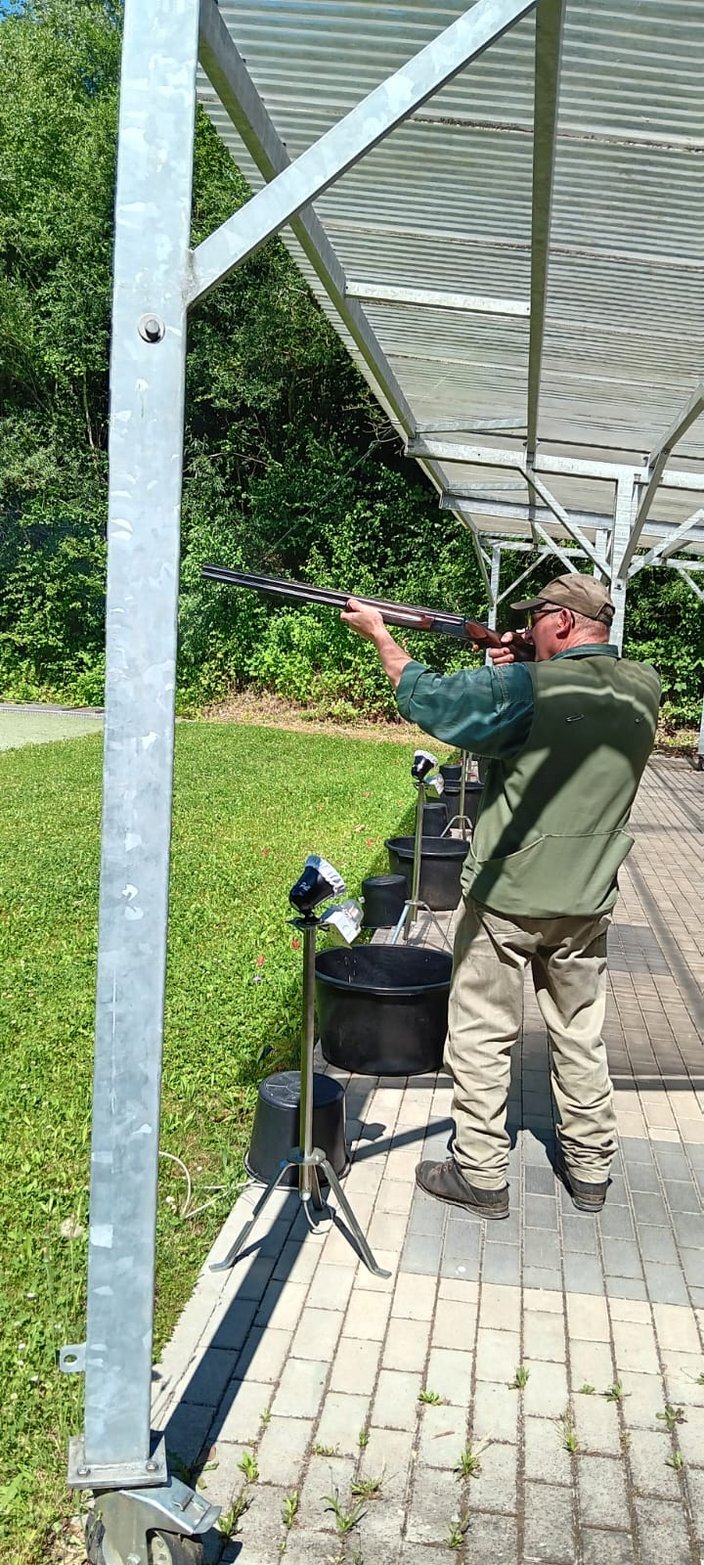 Ein Mann zielt mit einem Gewehr, steht unter einem überdachten Bereich draußen auf einer gepflasterten Fläche mit grüner Wiese im Hintergrund.