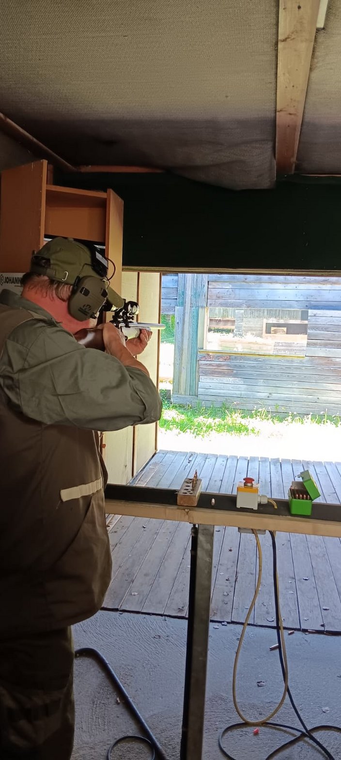 Eine Person zielt mit einem Gewehr auf einem Schießstand. Daneben steht ein Tisch mit Munition und einem gedrückten Knopf. Draußen sind Holzwände und Gras zu sehen.